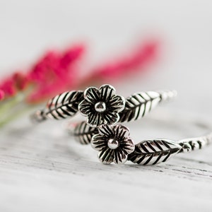 K&ouml;nnte beinhalten: Ein silberner Ring mit zwei Blumendesigns und Bl&auml;ttern. Der Ring liegt auf einer wei&szlig;en Holzoberfl&auml;che mit rosa Blumen im Hintergrund.