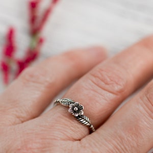 K&ouml;nnte beinhalten: Ein silberner Ring mit einem zarten Blumenmuster und Bl&auml;ttern. Der Ring wird an einem Finger getragen.