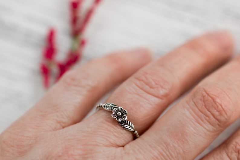K&ouml;nnte beinhalten: Ein silberner Ring mit einem zarten Blumenmuster und Bl&auml;ttern. Der Ring wird an einem Finger getragen.