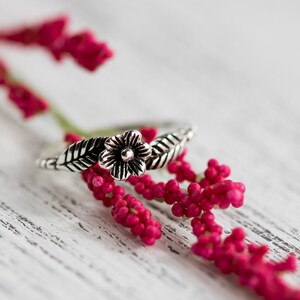 K&ouml;nnte beinhalten: Ein silberner Ring mit einem floralen Design mit einer Blume und Bl&auml;ttern. Der Ring liegt auf einem Zweig mit rosa Bl&uuml;ten.