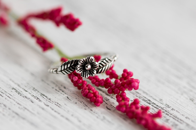 K&ouml;nnte beinhalten: Ein silberner Ring mit einem floralen Design mit einer Blume und Bl&auml;ttern. Der Ring liegt auf einem Zweig mit rosa Bl&uuml;ten.