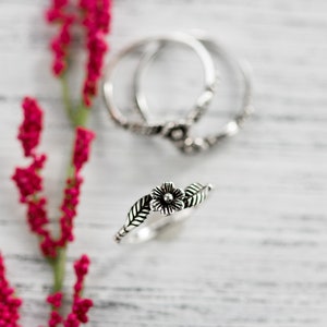 K&ouml;nnte beinhalten: Zwei silberne Ringe mit floralen Designs. Die Ringe sind &uuml;bereinander gestapelt. Der obere Ring hat eine einzelne Blume mit Bl&auml;ttern auf beiden Seiten. Der untere Ring hat ein filigraneres Blumenmuster.