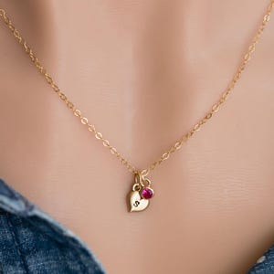 Puede incluir: Un collar de cadena de oro con un pequeño colgante de corazón y una sola piedra preciosa roja. El colgante de corazón tiene una letra 'S' grabada.