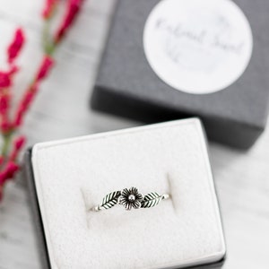 K&ouml;nnte beinhalten: Ein silberner Ring mit einem zarten Blumenmuster und zwei Bl&auml;ttern. Der Ring wird in einer wei&szlig;en Samt-Ringbox pr&auml;sentiert.
