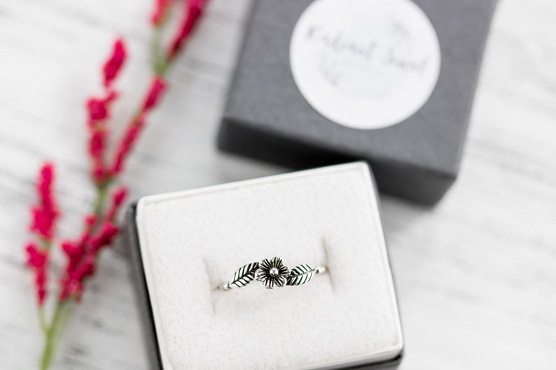 K&ouml;nnte beinhalten: Ein silberner Ring mit einem zarten Blumenmuster und zwei Bl&auml;ttern. Der Ring wird in einer wei&szlig;en Samt-Ringbox pr&auml;sentiert.