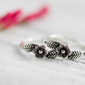 K&ouml;nnte beinhalten: Zwei silberne Ringe mit floralen Designs. Jeder Ring hat eine kleine Blume mit einem Punkt in der Mitte und zwei Bl&auml;ttern auf jeder Seite.