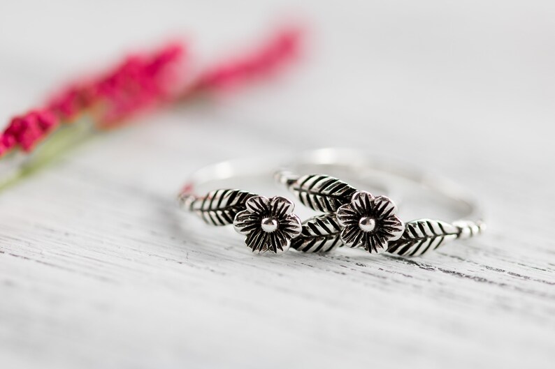 K&ouml;nnte beinhalten: Zwei silberne Ringe mit floralen Designs. Jeder Ring hat eine kleine Blume mit einem Punkt in der Mitte und zwei Bl&auml;ttern auf jeder Seite.
