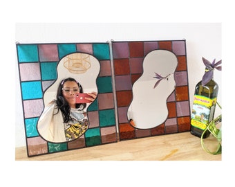 May include: Two stained glass mirrors with a unique, abstract shape. The mirror on the left has a blue and purple mosaic design, while the mirror on the right has a brown and purple mosaic design.