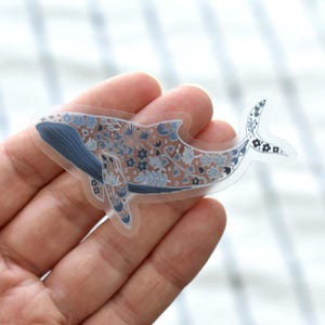 May include: A clear sticker featuring a whale design. The whale is decorated with blue and white floral patterns. The whale's body is a light brown color, and the fins are blue. The sticker is held in a hand.