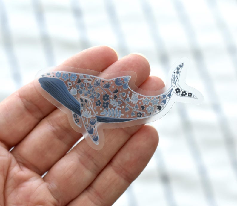 May include: A clear sticker featuring a whale design. The whale is decorated with blue and white floral patterns. The whale's body is a light brown color, and the fins are blue. The sticker is held in a hand.