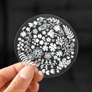 May include: A circular transparent sticker featuring a white line art illustration of various flowers, leaves, and botanical elements arranged in a dense, wreath-like pattern across the surface of the clear material.