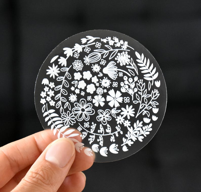 May include: A circular transparent sticker featuring a white line art illustration of various flowers, leaves, and botanical elements arranged in a dense, wreath-like pattern across the surface of the clear material.