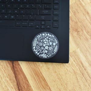 May include: A circular sticker featuring a white line-art botanical pattern of various flowers, leaves, and stems. The sticker is applied to the lower right corner of a black laptop keyboard, which has a standard QWERTY layout and black keys.