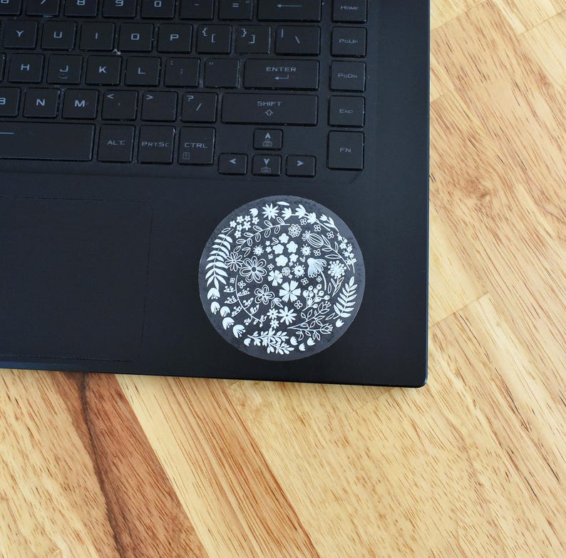 May include: A circular sticker featuring a white line-art botanical pattern of various flowers, leaves, and stems. The sticker is applied to the lower right corner of a black laptop keyboard, which has a standard QWERTY layout and black keys.