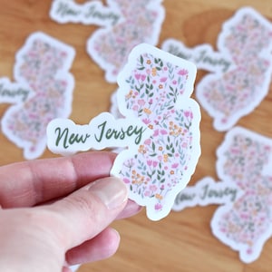 Può includere: Un adesivo fustellato a forma dello stato del New Jersey, caratterizzato da un motivo floreale con illustrazioni botaniche rosa, gialle e verdi. Il testo New Jersey è stampato in corsivo verde scuro al centro dell'adesivo.