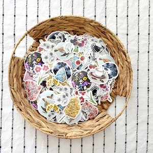 May include: A collection of stickers with floral, butterfly, and coffee designs, displayed in a woven basket. The stickers feature various colors and illustrations, including flowers, butterflies, and coffee cups. The basket sits on a white fabric with black vertical stripes.