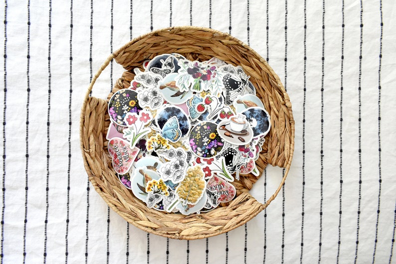 May include: A collection of stickers with floral, butterfly, and coffee designs, displayed in a woven basket. The stickers feature various colors and illustrations, including flowers, butterflies, and coffee cups. The basket sits on a white fabric with black vertical stripes.