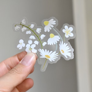 May include: A clear sticker featuring a floral design of white daisies and lily of the valley. The daisies have yellow centers and the lily of the valley has small white bell-shaped flowers. The sticker is held against a neutral background.