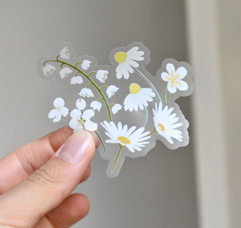 May include: A clear sticker featuring a floral design of white daisies and lily of the valley. The daisies have yellow centers and the lily of the valley has small white bell-shaped flowers. The sticker is held against a neutral background.