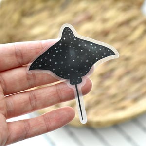 May include: A black and white sticker of a stingray with white spots. The sticker has a clear border and a thin, black tail. The sticker is held in a person's hand. The background is a blurred, woven texture.