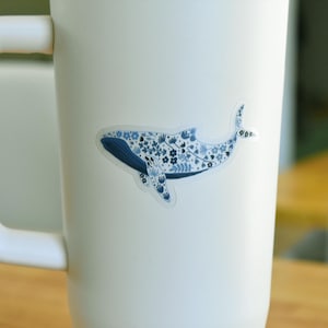 May include: A sticker of a whale with a blue floral pattern, affixed to a white mug. The whale design features a dark blue body and tail, with a lighter blue floral pattern filling the body.