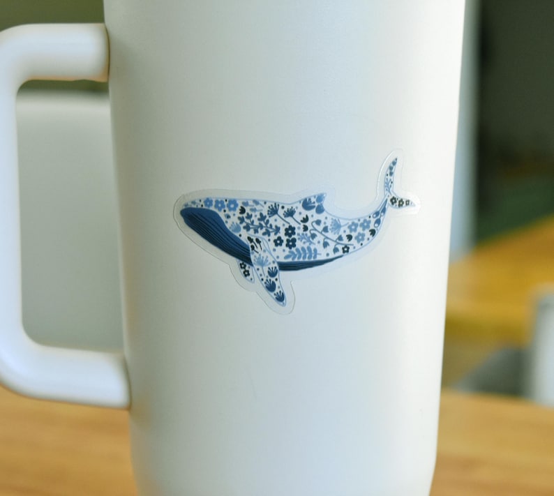 May include: A sticker of a whale with a blue floral pattern, affixed to a white mug. The whale design features a dark blue body and tail, with a lighter blue floral pattern filling the body.