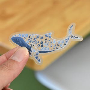 May include: A whale sticker with a floral design. The whale is blue and beige, with blue floral patterns. The sticker is held by a hand against a wooden background.