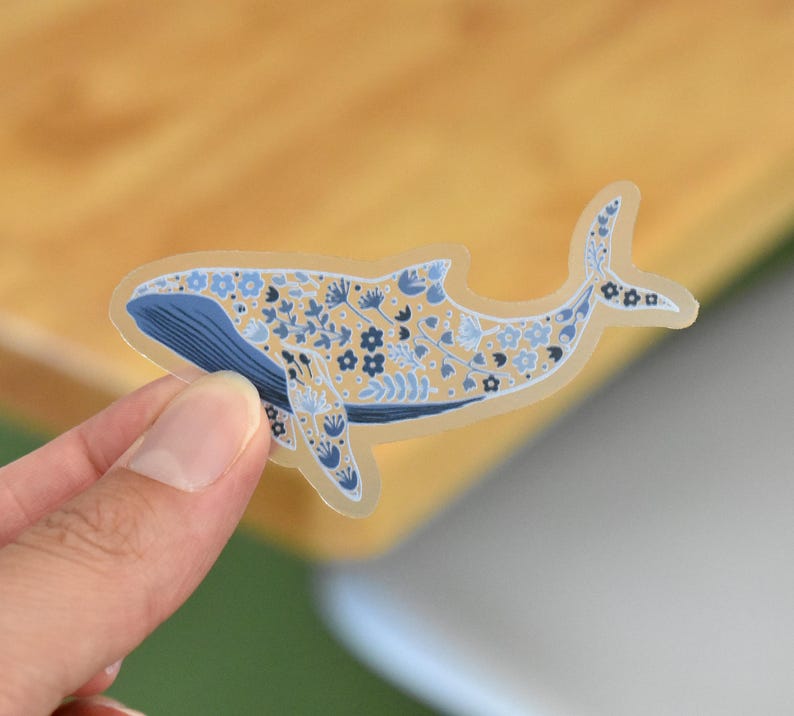 May include: A whale sticker with a floral design. The whale is blue and beige, with blue floral patterns. The sticker is held by a hand against a wooden background.