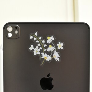 May include: A clear sticker featuring a floral design of white daisies and lily of the valley flowers with green stems. The sticker is placed on the back of a black tablet, next to a white stylus.