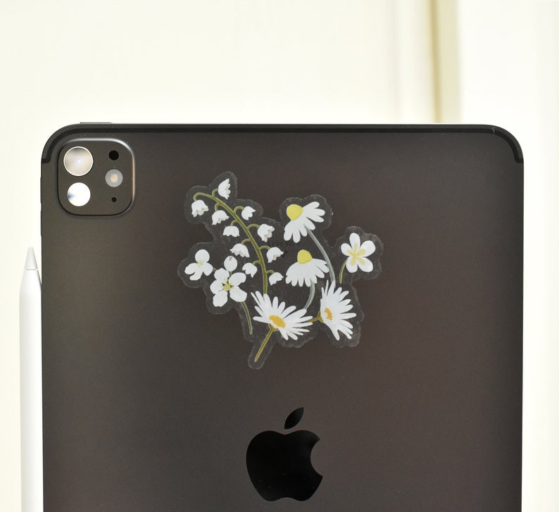 May include: A clear sticker featuring a floral design of white daisies and lily of the valley flowers with green stems. The sticker is placed on the back of a black tablet, next to a white stylus.