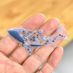 May include: A clear sticker featuring a whale design. The whale is blue with a floral pattern in shades of blue, white, and tan. The sticker is held in a hand.