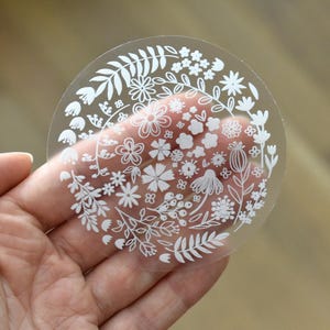 May include: A circular, transparent sticker featuring a white line-art illustration of various flowers, leaves, and botanical elements arranged in a dense, wreath-like pattern across the surface of the clear material.