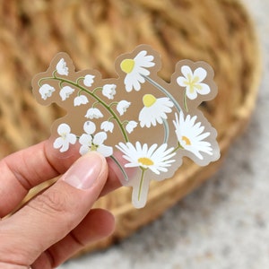 May include: A clear sticker featuring a floral design of white daisies and lily of the valley flowers with yellow centers and green stems. The sticker has a transparent background and is held by a hand.