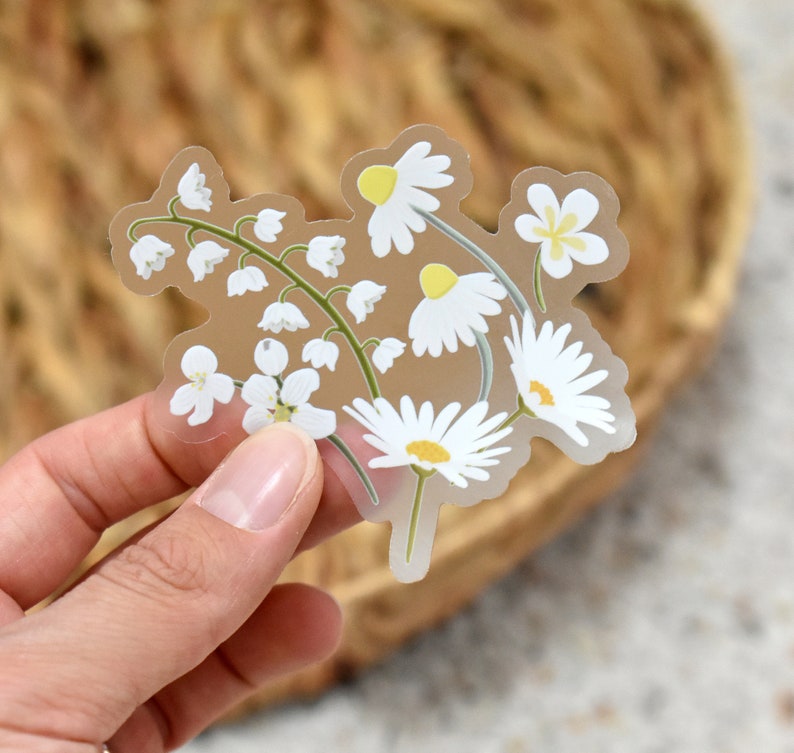 May include: A clear sticker featuring a floral design of white daisies and lily of the valley flowers with yellow centers and green stems. The sticker has a transparent background and is held by a hand.