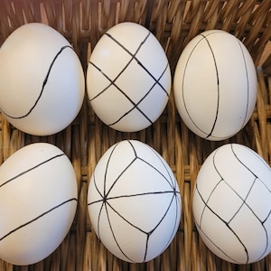 Puede incluir: Seis huevos blancos con diseños geométricos negros, incluyendo líneas y sombreado cruzado, están dispuestos en una cesta tejida. Los diseños de los huevos varían, creando una estética moderna y artística. La textura de la cesta añade un contraste rústico.