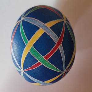 May include: A blue decorative egg with a colorful woven pattern. The egg features intersecting bands of red, yellow, green, and light blue, creating a geometric design. The egg is a unique decorative item.