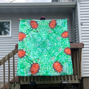 May include: A large green and orange tie-dye tapestry with a circular pattern. The tapestry is being held up by a person.