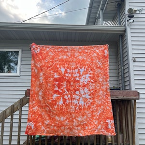 May include: Orange and white tie-dye fabric hanging on a wooden deck railing. The fabric has a circular pattern with a white center and orange radiating outwards.