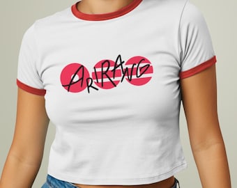 May include: White crop top with red trim and the word "ARRANG" in black script. The word is centered over three red circles. The model is wearing blue jeans and smiling.