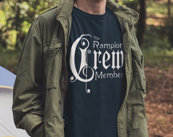 May include: A man wearing a dark blue t-shirt with a white graphic that says "The Rampion Crew Member" and a green jacket. He is standing in a forest with a tent behind him.