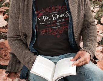 May include: A man wearing a black t-shirt with a red graphic that says "The Dregs" and the text "When you can't beat the odds, change the game." He is wearing a grey beanie and a brown cardigan. He is sitting on the ground with a book in his lap.