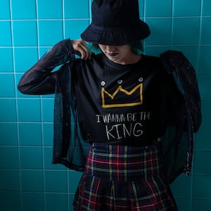 May include: A person wearing a black t-shirt with a yellow crown and the text "I wanna be the KING" in white. They are also wearing a black and red plaid skirt and a black bucket hat.