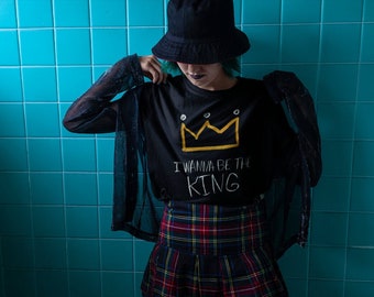 May include: A person wearing a black t-shirt with a yellow crown and the text "I wanna be the KING" in white. They are also wearing a black and red plaid skirt and a black bucket hat.