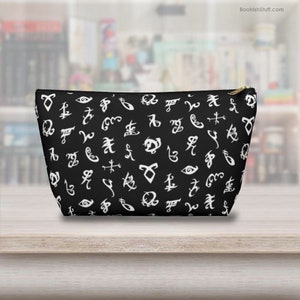 May include: Black zippered pouch with a white pattern of runes and symbols. The pouch is made of fabric and has a zipper closure.