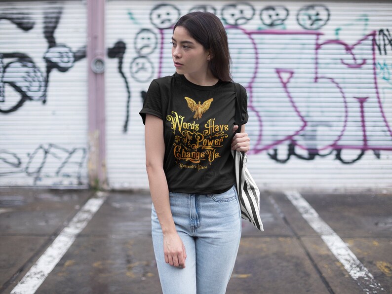 Puede incluir: Camiseta negra con un gr&aacute;fico dorado de un p&aacute;jaro y el texto "Words Have The Power To Change Us." El texto est&aacute; en una fuente vintage.