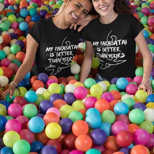 May include: Two people wearing black t-shirts with white text that says "My parabatai is better than yours" and a drawing of a parabatai symbol. They are sitting in a ball pit filled with colorful balls.