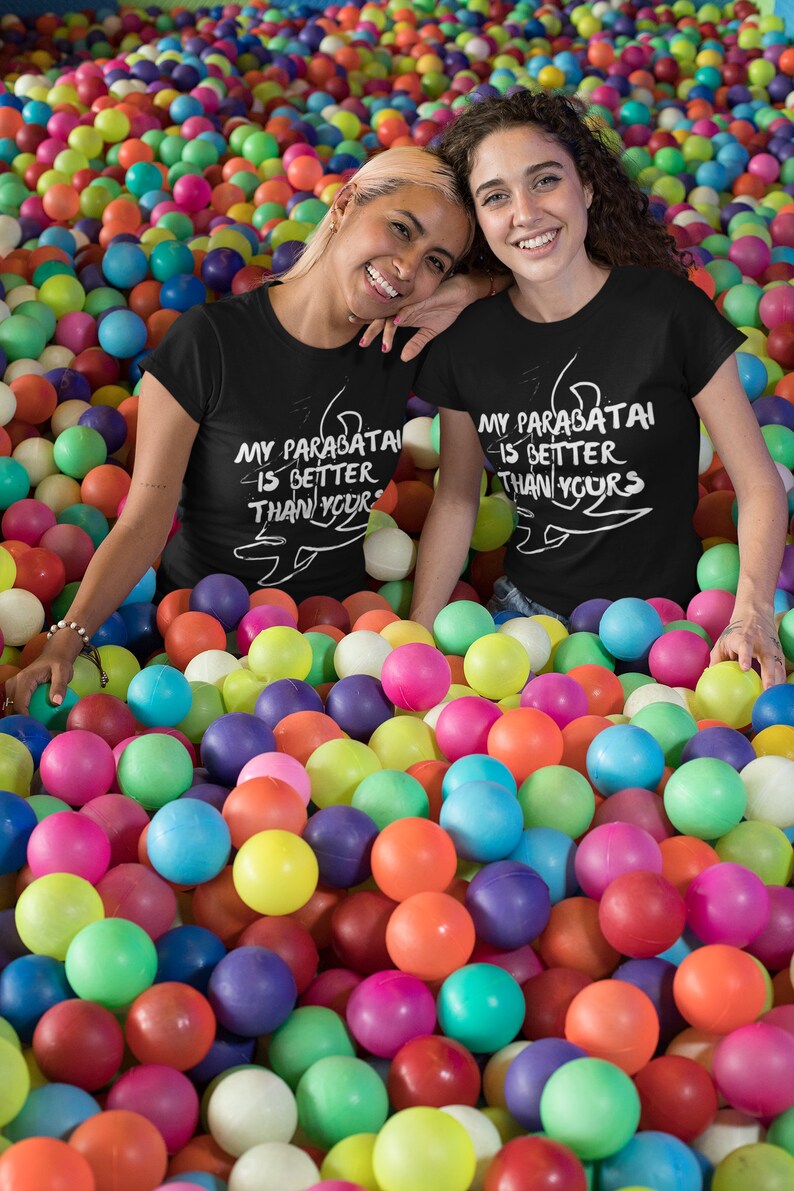 May include: Two people wearing black t-shirts with white text that says "My parabatai is better than yours" and a drawing of a parabatai symbol. They are sitting in a ball pit filled with colorful balls.