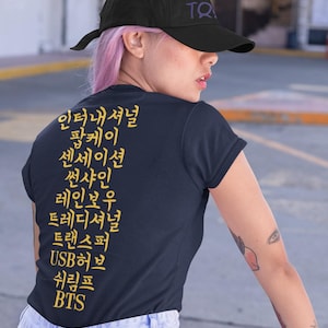Könnte beinhalten: Ein marineblaues T-Shirt mit koreanischem Text in Gelb. Der Text lautet "International Pop Key, Sensation, Sunshine, Rainbow, Traditional, Transfer, USB Hub, Shrimp, BTS". Das Shirt wird von einer Person getragen, die eine schwarze Baseballkappe mit dem Text "BT MAP OF THE SOUL" in Lila trägt.