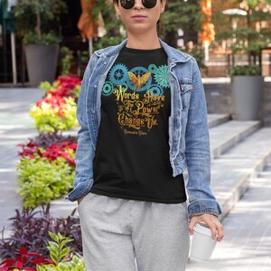 Puede incluir: Una camiseta negra con un dise&ntilde;o gr&aacute;fico de un p&aacute;jaro dorado con las alas extendidas, rodeado de engranajes azules y verdes. El texto "Words Have The Power To Change Us." est&aacute; impreso debajo del p&aacute;jaro. La cita se atribuye a "Cassandra Clare".