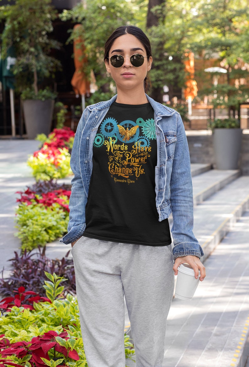 Puede incluir: Una camiseta negra con un dise&ntilde;o gr&aacute;fico de un p&aacute;jaro dorado con las alas extendidas, rodeado de engranajes azules y verdes. El texto "Words Have The Power To Change Us." est&aacute; impreso debajo del p&aacute;jaro. La cita se atribuye a "Cassandra Clare".